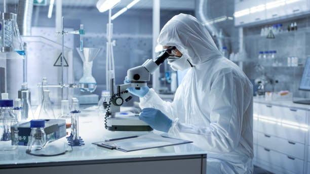 in a secure high level research laboratory scientist in a coverall examines petri dish under microscope. - protective suit fotos imagens e fotografias de stock