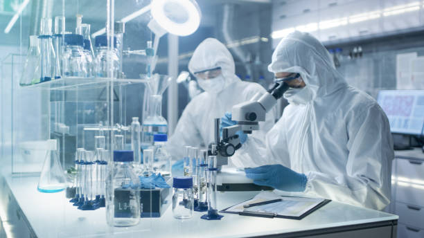 in a secure high level laboratory scientists in a coverall conducting a research. chemist adjusts samples in a  petri dish with pincers and then examines them under microscope and his colleague writes down results. - smiling research science and technology clothing imagens e fotografias de stock