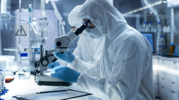 istock In a Secure High Level Laboratory Scientists in a Coverall Conducting a Research. Chemist Examines Samples Under Microscope and His Colleague Writes Down Results. 823447820