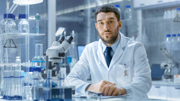 young male research scientist talks into camera. he's sitting in a high-end modern laboratory with beakers, glassware, microscope and working monitors surround him. - smiling research science and technology clothing imagens e fotografias de stock