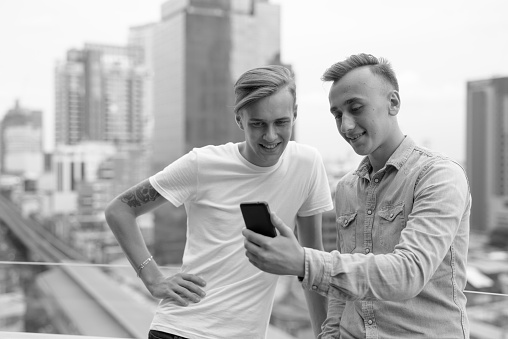 Portrait of two young handsome teenage boys using phone together against view of the city in Bangkok Thailand in black and white horizontal shot