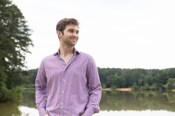 A millennial man in his late 20s stands in front of a lake.  He is smiling and looking off camera.  He is caucasian, has a light beard, dark brown hair, and wears a purple gingham shirt.