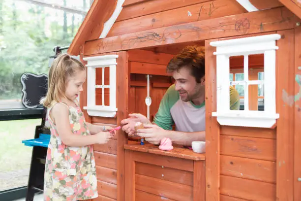 A father and daughter play with a playhouse.  The dad sits inside the tiny house and interacts with his daughter through a window.  They are playing with plastic food toys.  They are caucasian, the dad has a light beard and dark brown hair, the daughter has pigtails and light brown hair.