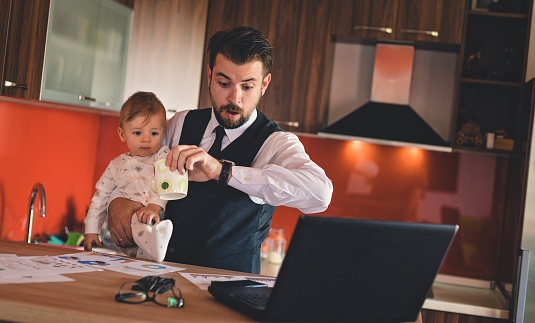 Single businessman father checking the time while holding baby boy in the kitchen