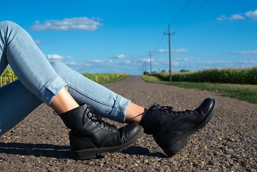 Boots and jeans of young woman hitchhiker sitting in the middle of the road on sunny summer day.