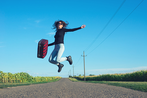 Young woman hitchhiker jumping high in the air in the middle of the road