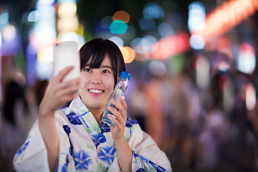 A young Japanese woman is in Yukata, taking a selfie with a smartphone and holding ramine during a traditional festival in Tokyo.