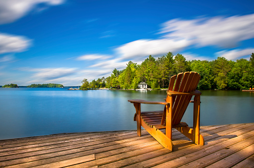 Adirondack chair on a wood dock facing a calm lake. Across the water is a white cottage nestled among green trees. There is a boat dock on the water in front of the cottage.