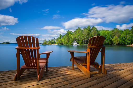 Two Muskoka chairs sitting on a wood dock facing a ake. Across the calm water is a white cottage nestled among green trees. There is a boat dock on the water in front of the cottage.