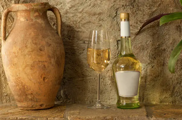 an old clay jug and a bottle of white wine with a wineglass stand on the background of a clay wall