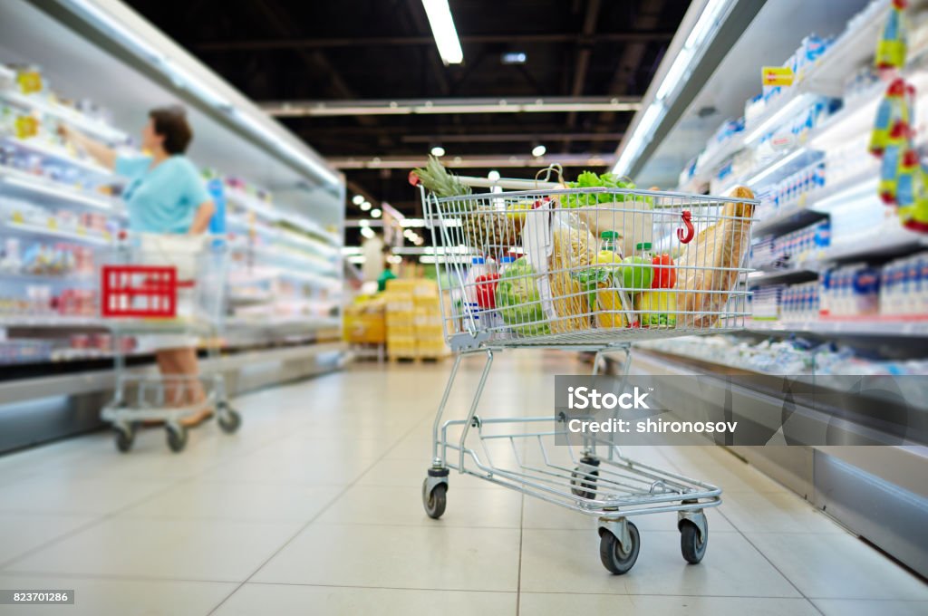 Shopping cart with products Shopping cart full of grocery standing next to shelves with dairy products in hypermarket Shopping Cart Stock Photo