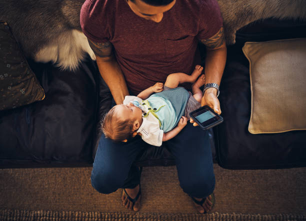 Distracted from daddy duties Shot of a young man using a mobile phone while holding his newborn baby boy at home ignoring stock pictures, royalty-free photos & images