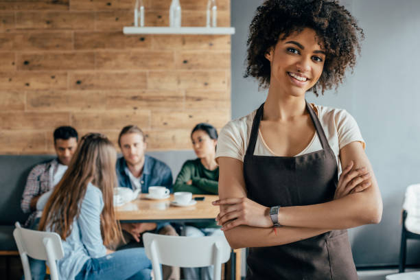 Smiling african american waitress standing with customers sitting behind in cafe Smiling african american waitress standing with customers sitting behind in cafe waitress stock pictures, royalty-free photos & images
