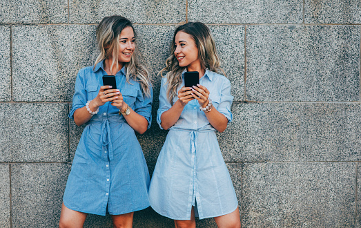 Twins using smart phones in front of a wall outdoors.