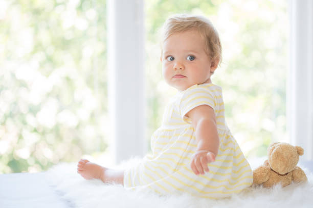 Adorable baby girl sitting on fluffy blanket in front of window, looking over shoulder. Wearing yellow dress and holding teddy bear.
