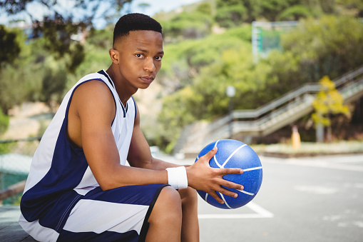 Portrait of teenage boy with basketball sitting on bench in court