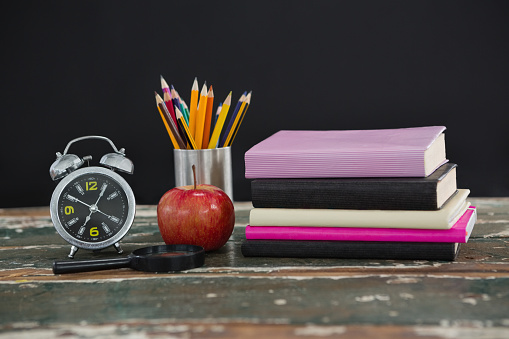 Stack of books with alarm clock, magnifying glass, apple and pen holder on wooden table