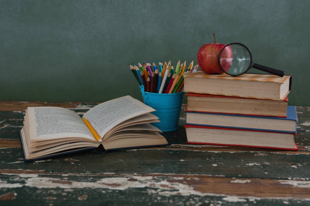 Stack of books with magnifying glass, apple and pen holder on wooden table