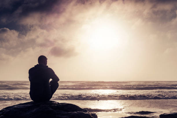 istock Man sitting Contemplating on a rock, Watergate Bay Beach, Sunset. 823917916