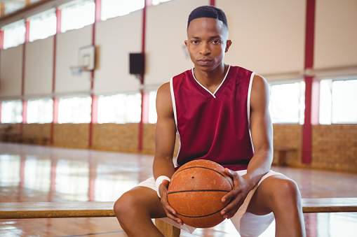 Portrait of confident male teenager with basketball sitting on bench in court