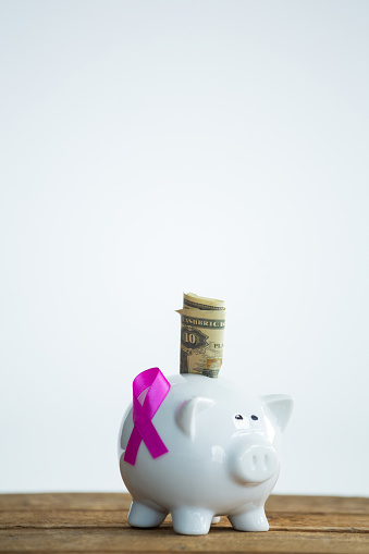 Paper currency and pink Breast Cancer Awareness ribbon on piggybank over wooden table against white background