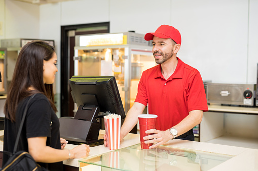Portrait of a good looking worker in a concession food in a movie theater serving popcorn and soda to a female customer