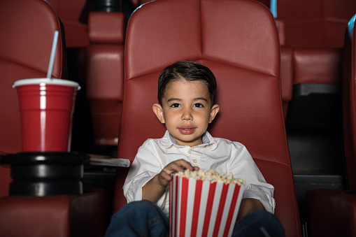 Portrait of a good looking Hispanic boy sitting in a movie theater and eating a lot of popcorn