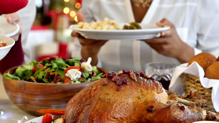 Friends and family gather to celebrate Thanksgiving or Christmas. They are enjoying a traditional holiday dinner or lunch. Handheld camera pans across a table filled with a variety of foods including macaroni and cheese, pie, a basketful of dinner rolls, a turkey with all of the trimmings and a salad. A candle is burning on the table. The unrecognizable female party host serves her guest.