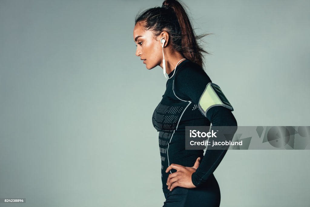 Fit young woman standing with hands on hips Side view of young woman listening to music with earphones. Fit female model standing with her hands on hips against grey background. Exercising Stock Photo