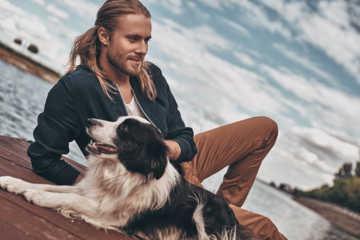 Handsome young man and his dog sitting near the lake while spending time outdoors