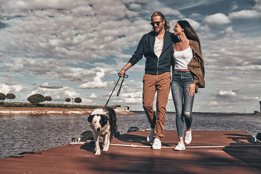 Full length of beautiful young couple walking with their dog on the river shore while spending time outdoors