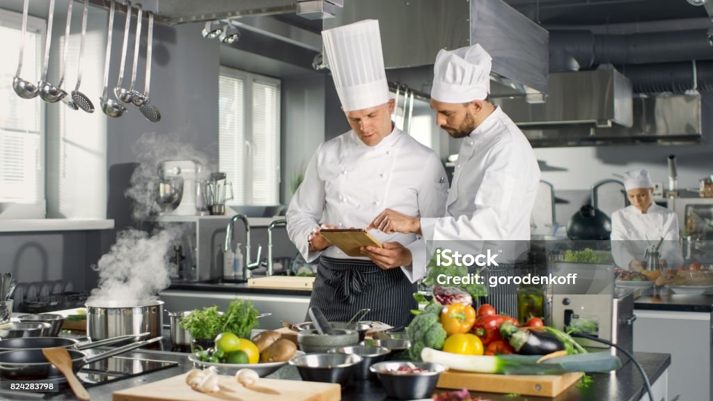 Dos Chefs famosos discutan su vídeo Blog mientras uso de Tablet PC. Trabajan en una cocina profesional grande restaurante de acero inoxidable. - Foto de stock de Chef libre de derechos