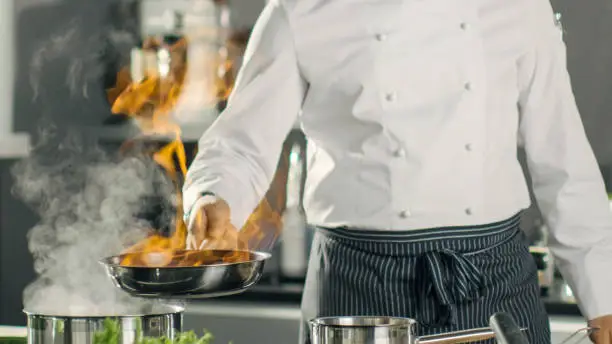 Photo of Close-up Shot of Fireing Oil on a Pan. Flambe Style Cooking. Cook Works in a Modern Kitchen with Lots of Ingredients are Around.