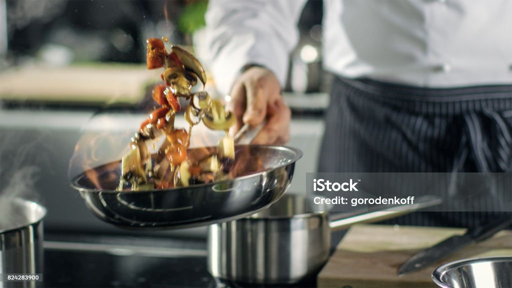 Professional Chef Cooks Flambe Style. He Prepares Dish in a Pan with Open Flames. He Works in a Modern Kitchen with Different Ingredients Lying Around. Chef Stock Photo
