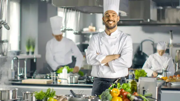 Photo of Famous Chef of a Big Restaurant Crosses Arms and Smiles in a Modern Kitchen. His Staff in Working in the Background.