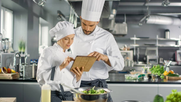 istock Male and Female Famous Chefs Discuss Their Video Blog while Using Tablet Computer. They Work on a Big Restaurant Stainless Steel Professional Kitchen. 824284062