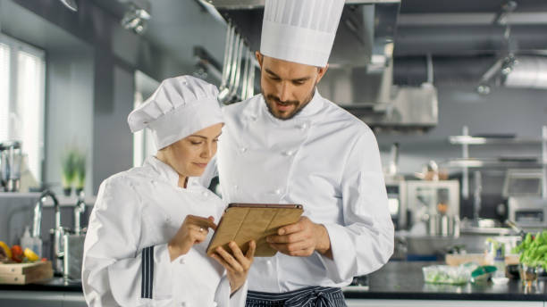 istock Male and Female Famous Chefs Discuss Their Video Blog while Using Tablet Computer. They Work on a Big Restaurant Stainless Steel Professional Kitchen. 824284074
