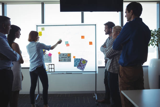 istock Businesswoman sticking adhesive notes on whiteboard while colleagues standing at office 824563388
