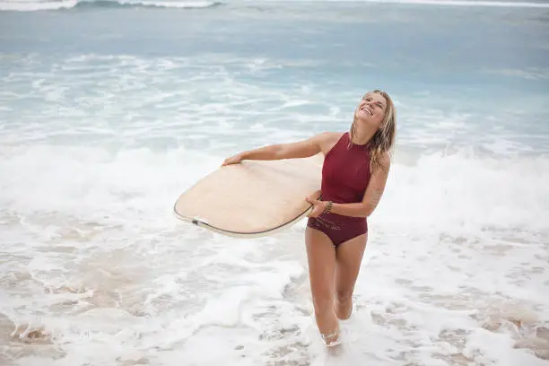 Beautiful smiling surf girl in dark-red swimming suit is going out of the ocean with a surfboard