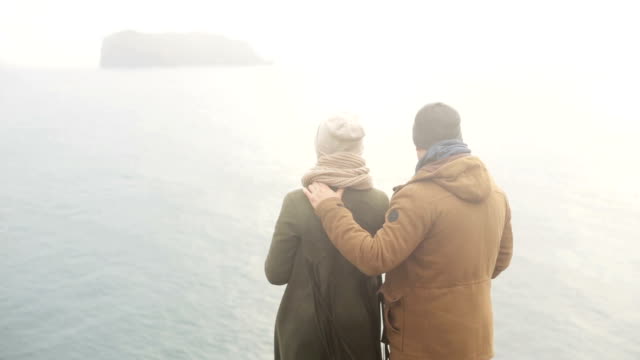 Back view of young man and woman standing on the shore of the sea and enjoying the beautiful view in cold foggy day
