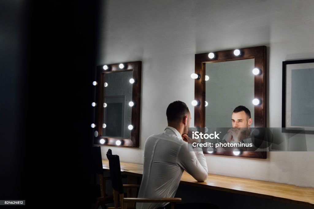 Portrait of handsome young man in white shirt looking into the mirror Portrait of handsome young man in white shirt looking into the mirror. Male reflection, light bulbs. Dressing Room Stock Photo