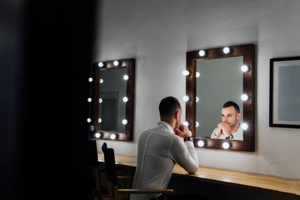 Portrait of handsome young man in white shirt looking into the mirror Portrait of handsome young man in white shirt looking into the mirror. Male reflection, light bulbs. backstage mirror stock pictures, royalty-free photos & images