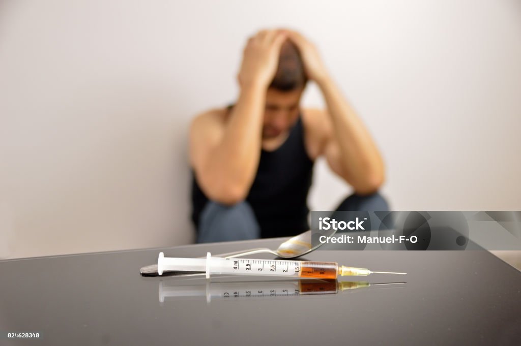 drug addict to heroin cropped shot of junkie sitting in the background with a close-up of syringe and small spoon with cooked heroine on the table Addict Stock Photo