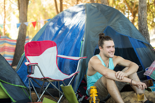 Thoughtful young man holding beer glass while sitting by tent at campsite