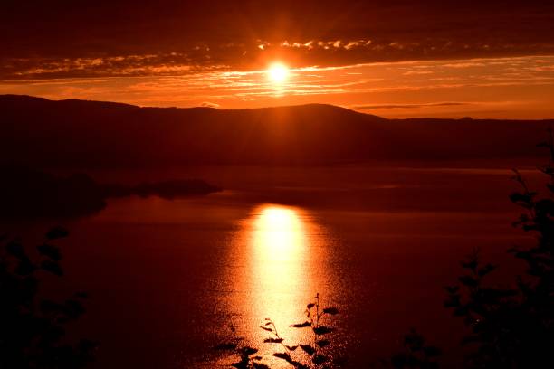 Silhouettes of a forest at the shore of a river with a reflection of the midnight sun, shades of yellow, blue and gold in Finnish Lapland.