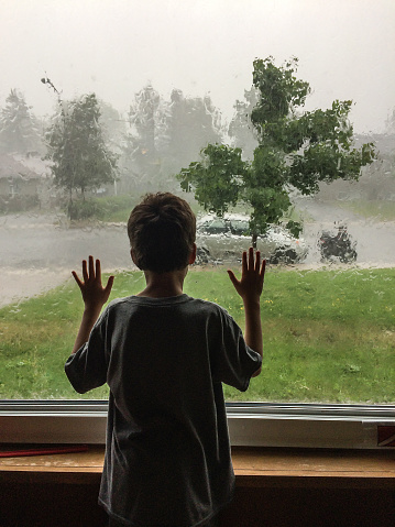 Litlle boy seen from behind and looking at the rain storm through living room window