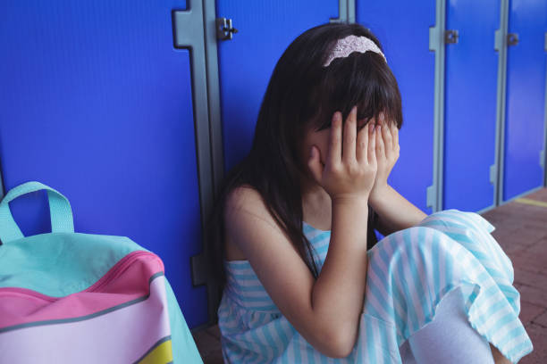 istock Schoolgirl covering face with hands while sitting by lockers 824697190