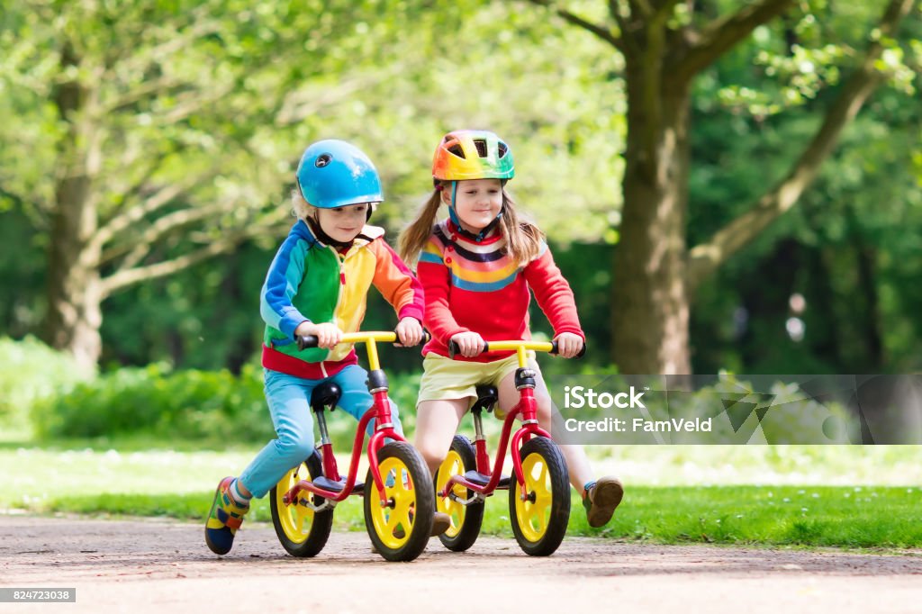 Kids ride balance bike in park Children riding balance bike. Kids on bicycle in sunny park. Little girl and boy ride glider bike on warm summer day. Preschooler learning to balance on run bicycle in safe helmet. Sport for kids. Cycling Stock Photo