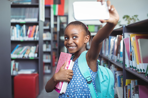 Smiling girl taking selfie while standing by bookshelf in library