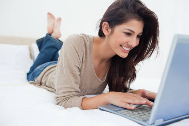 femme couchée au lit avec son ordinateur portable devant elle et souriant - using computer bedroom one woman only cheerful photos et images de collection